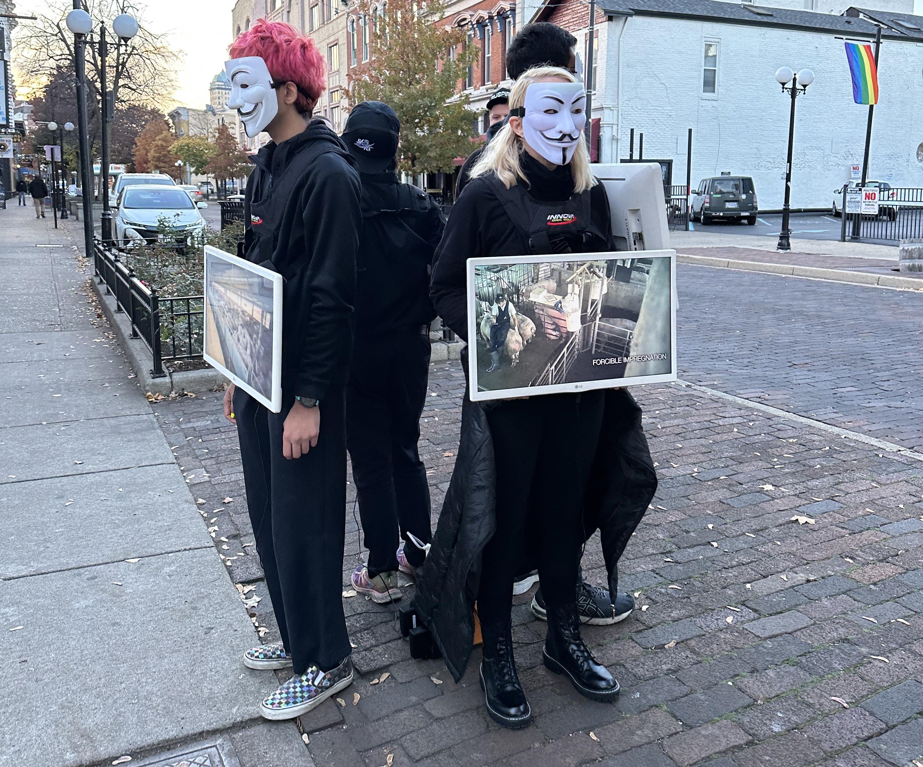 People standing in a cube facing outward holding TVs with Guy Fox masks